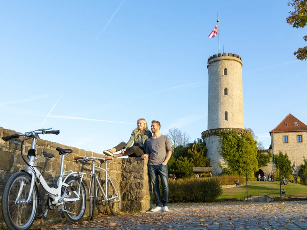 Bielefelder Romanzen_Sparrenburg_Ulrich Helweg.jpg Zwei Personen sitzen auf einer Mauer vor einem mittelalterlichen Turm und genießen die Herbstlandschaft.