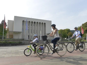 Bielefelder Silhouetten Nord_Rudolf-Oetker-Halle_Ulrich Helweg.jpg Familie mit Helmen fährt fröhlich Rad auf einem breiten Weg vor einem modernen, weißen Gebäude.