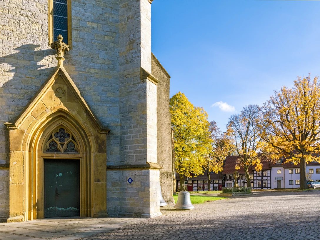Bielefelder Silhouetten Nord_Stiftskirche Schildesche_Ulrich Helweg.jpg Eingang einer historischen Kirche mit Herbstbäumen und Fachwerkhäusern im Hintergrund.