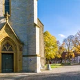 Bielefelder Silhouetten Nord_Stiftskirche Schildesche_Ulrich Helweg.jpg Eingang einer historischen Kirche mit Herbstbäumen und Fachwerkhäusern im Hintergrund.