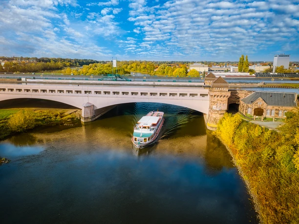 Minden-Wasserstraßenkreuz-Teutoburger-Wald-Tourismus-J-Motzny-001-CC-BY-SA.jpg Ein Ausflugsschiff durchquert den Mittellandkanal in Minden, mit einer Brücke und Herbstlaub im Hintergrund.