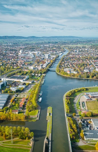 Minden-Wasserstraßenkreuz-Teutoburger-Wald-Tourismus-J-Motzny-092.jpg Luftaufnahme von Minden mit Weser und Industriegebiet im Vordergrund, umgeben von grüner Landschaft.