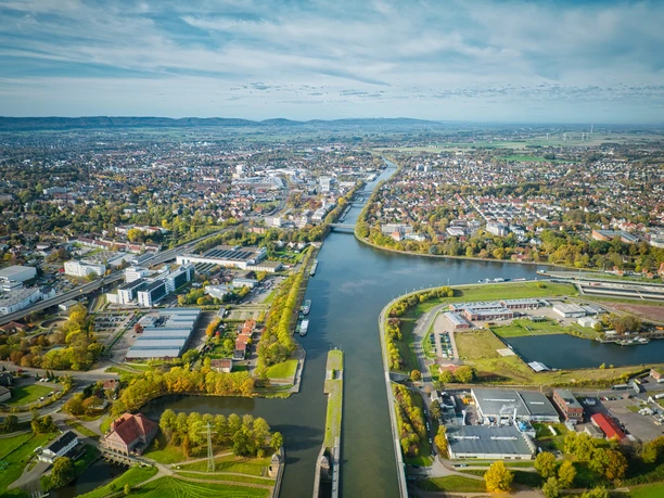 Minden-Wasserstraßenkreuz-Teutoburger-Wald-Tourismus-J-Motzny-092.jpg Luftaufnahme von Minden mit Weser und Industriegebiet im Vordergrund, umgeben von grüner Landschaft.