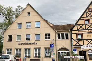 Two-storey building with the inscription "Tourist Information", next to it half-timbered house with restaurant "Elbterrasse", cloudy sky.