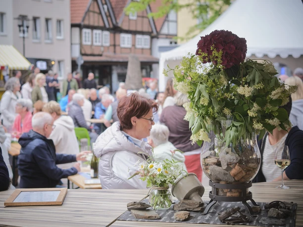 Eine festliche Versammlung bei einem Weinfest im Freien, mit Menschen an Tischen und Blumenarrangement.