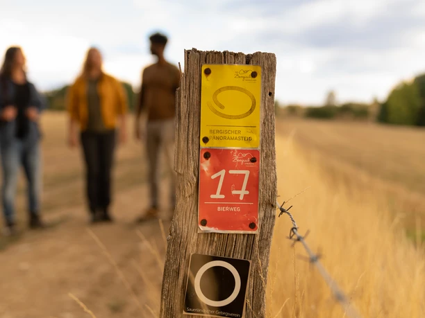 Nahaufnahme eines Holzzauns mit Wanderwegschildern; unscharfe Wanderer im Hintergrund auf einem Pfad.