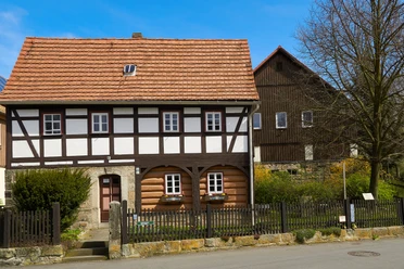 Heimatstube Reinhardsdorf Schöna Fachwerkhaus mit rotem Ziegeldach und weißen Fenstern, umgeben von einem Holzzaun und einem blühenden Garten, bei sonnigem Wetter.