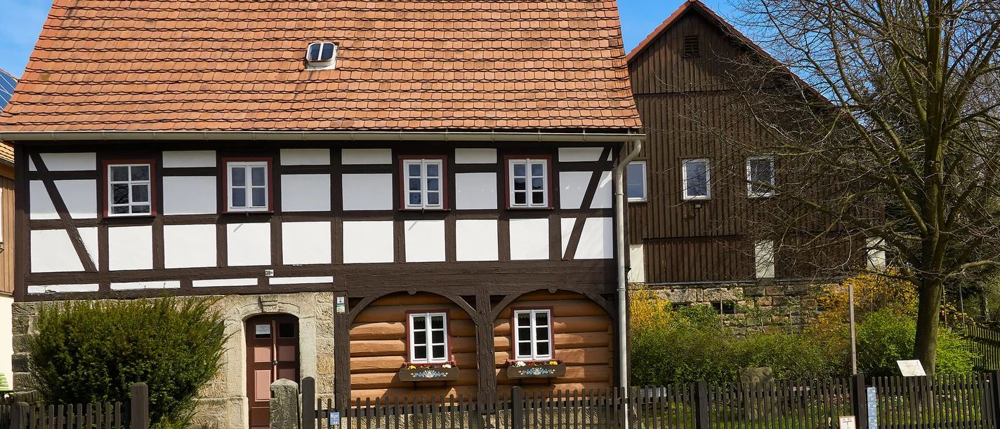 Heimatstube Reinhardsdorf Schöna Fachwerkhaus mit rotem Ziegeldach und weißen Fenstern, umgeben von einem Holzzaun und einem blühenden Garten, bei sonnigem Wetter.