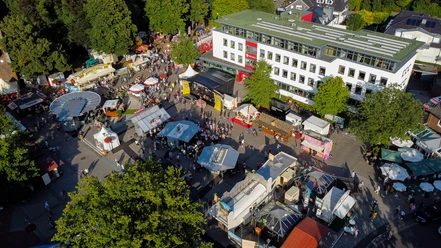 bad-zwischenahner-woche-marktplatz-rathaus-von-oben.jpg Auf dem Bild ist der Marktplatz von Bad Zwischenahn während eines Festes, gefüllt mit bunten Ständen und Menschen. Das Rathaus ragt links empor, umgeben von üppigem Grün, das eine lebendige Atmosphäre schafft.