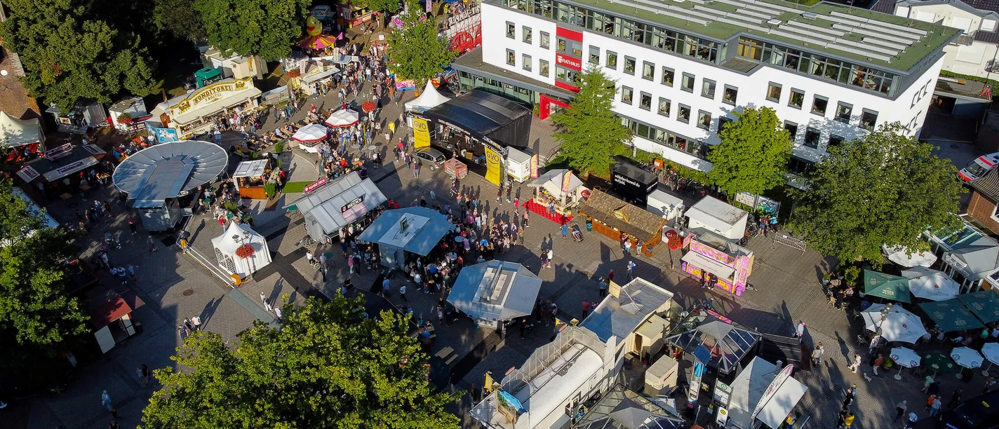bad-zwischenahner-woche-marktplatz-rathaus-von-oben.jpg The photo shows the town square in Bad Zwischenahn during a festival, filled with colorful stalls and people. The town hall towers above on the left, surrounded by lush greenery that creates a lively atmosphere.