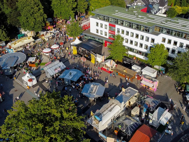 bad-zwischenahner-woche-marktplatz-rathaus-von-oben.jpg Auf dem Bild ist der Marktplatz von Bad Zwischenahn während eines Festes, gefüllt mit bunten Ständen und Menschen. Das Rathaus ragt links empor, umgeben von üppigem Grün, das eine lebendige Atmosphäre schafft.