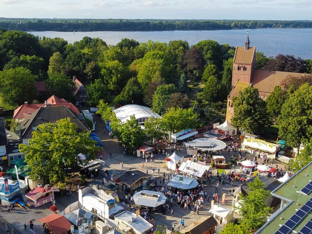 bad-zwischenahner-woche-von-oben-kirche-meer.jpg Die Luftaufnahme zeigt ein buntes Stadtfest um eine Kirche, eingebettet in viel Grün am Seeufer.