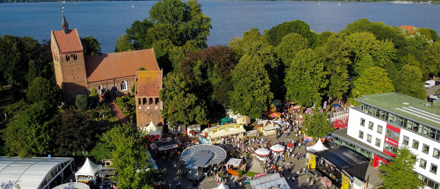 Luftaufnahme eines belebten Marktgeländes in Bad Zwischenahn mit Kirche im Hintergrund und See im Westen.
