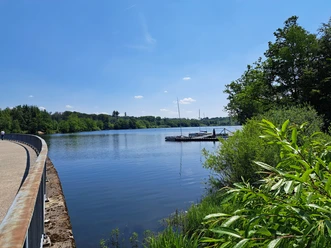 Bergischer Fuhrmannsweg Talsperre an einem sonnigen Tag, mit Steg und Booten, umgeben von grüner Natur und blauem Himmel.