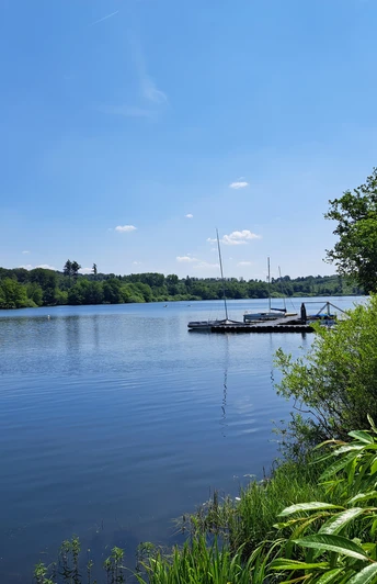 Bergischer Fuhrmannsweg Talsperre an einem sonnigen Tag, mit Steg und Booten, umgeben von grüner Natur und blauem Himmel.