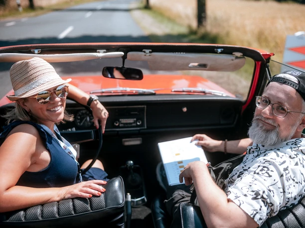PS.Rallye 2023 Zwei Personen sitzen in einem roten Oldtimer bei sonnigem Wetter.