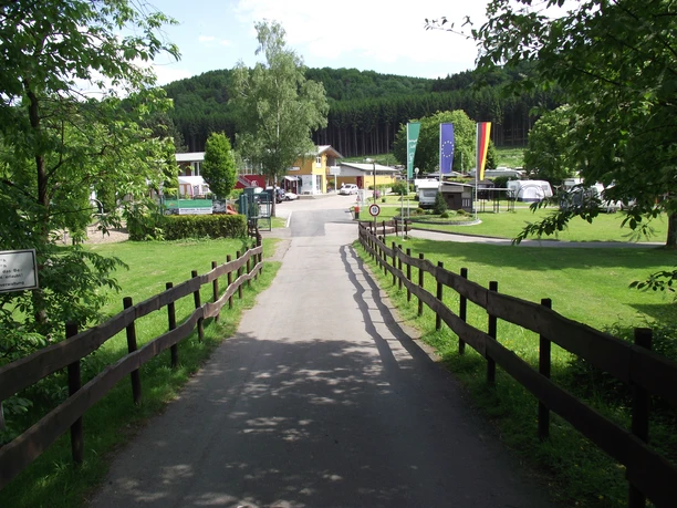 DSCF2699 Kopie.jpg Weg führt durch grüne, von Holzgeländern gesäumte Felder zu einem Campingplatz mit bunten Fahnen.