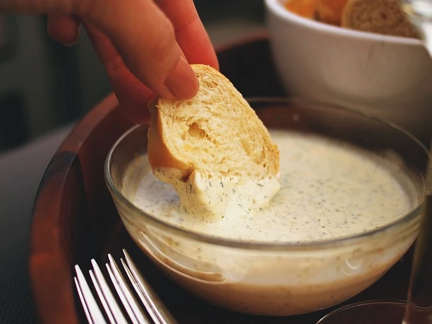 Beipielbild Eine Hand taucht frisches Baguette in eine Schale mit cremigem Dip neben Salatgabeln.