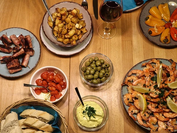 Beipielbild Vielfältige Tapasauswahl mit gegrilltem Fleisch, Garnelen, buntem Gemüse, Brot, Oliven und Wein.