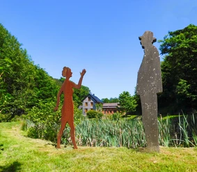 Sinneswald-Braun-Brudes.jpg Zwei Skulpturen, eine Frau und ein Mann, stehen auf einer grünen Wiese vor einem Haus im Hintergrund.