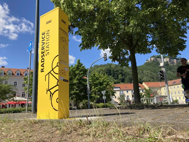 ADAC Fahrradstation Königstein Gelbe Radservicestation des ADAC neben einem Baum, im Hintergrund Gebäude mit roten Dächern und ein bewaldeter Hügel unter blauem Himmel.