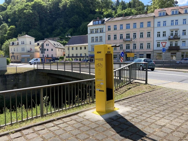 ADAC Fahrradstation Königstein Gelbe ADAC-Radservicestation neben einer Straße mit mehrstöckigen Häusern im Hintergrund, umgeben von grünen Bäumen und sonniger Atmosphäre.