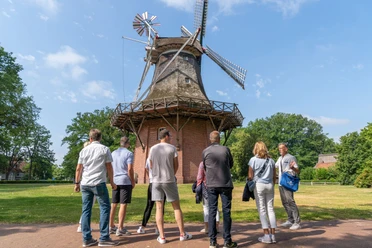 mühle.jpg Eine Gruppe von Menschen betrachtet eine historische Windmühle bei sonnigem Wetter im Grünen.