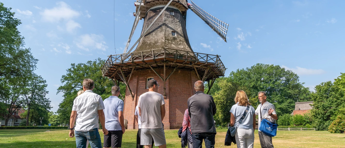 mühle.jpg Eine Gruppe von Menschen betrachtet eine historische Windmühle bei sonnigem Wetter im Grünen.