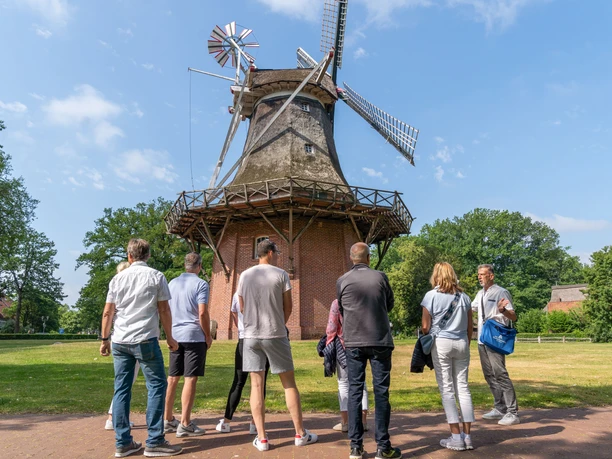 mühle.jpg Eine Gruppe von Menschen betrachtet eine historische Windmühle bei sonnigem Wetter im Grünen.
