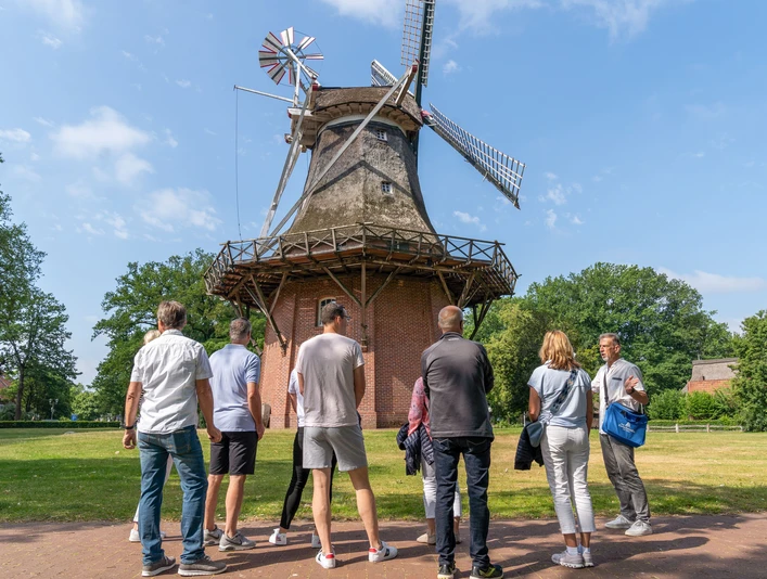 mühle.jpg Eine Gruppe von Menschen betrachtet eine historische Windmühle bei sonnigem Wetter im Grünen.