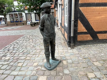 Calenberger Bur Bronzefigur eines Mannes mit Hut in traditioneller Kleidung, stehend auf einem gepflasterten Platz.