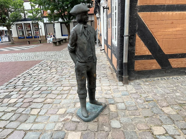 Calenberger Bur Bronzefigur eines Mannes mit Hut in traditioneller Kleidung, stehend auf einem gepflasterten Platz.