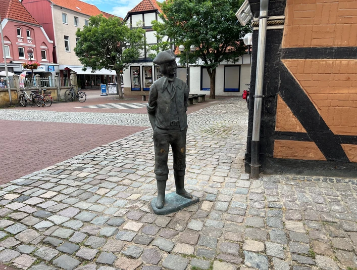 Bronzefigur eines historischen Arbeiters mit Schlägermütze auf gepflastertem Platz in Stadtkulisse.