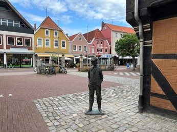Calenberger Bur Eine bronzene Jungenfigur steht auf einem Platz vor bunten Häusern und einem Café im Hintergrund.