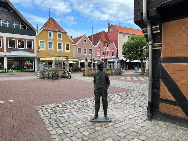 Calenberger Bur Eine bronzene Jungenfigur steht auf einem Platz vor bunten Häusern und einem Café im Hintergrund.