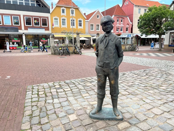 Ein bronzener Junge in traditioneller Kleidung steht auf einem gepflasterten Platz vor bunten Häusern.