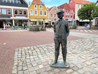 Calenberger Bur Ein bronzener Junge in traditioneller Kleidung steht auf einem gepflasterten Platz vor bunten Häusern.