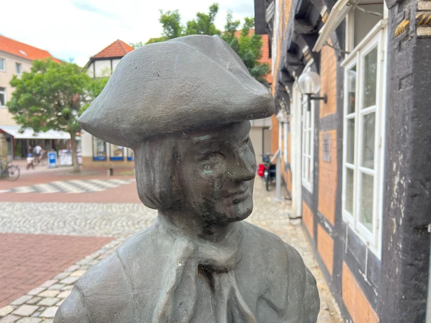 Bronzestatue eines Mannes mit Hut und Halstuch, im Hintergrund Fachwerkhäuser und gepflasterte Gassen.