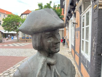 Calenberger Bur Bronzestatue eines Mannes mit Hut und Halstuch, im Hintergrund Fachwerkhäuser und gepflasterte Gassen.