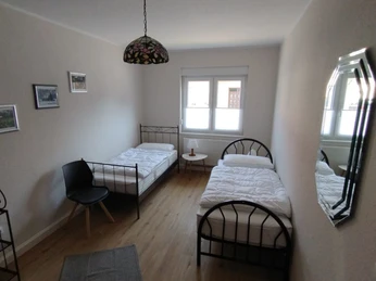 Burkis Ferienhaus Zwei Einzelbetten in einem schlichten, hellen Raum mit Parkettboden und Wandbildern, Tageslicht erhellt.Two single beds in a simple, bright room with parquet flooring and murals, illuminated by natural light.To enkeltsenge i et enkelt, lyst værelse med parketgulv og vægmalerier, oplyst af naturligt lys.Twee eenpersoonsbedden in een eenvoudige, lichte kamer met parketvloer en muurschilderingen, verlicht door daglicht.