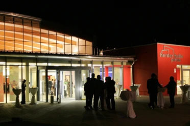 Buitenaanzicht van de Herderhalle Pirna bij nacht, verlicht met mensen die voor de ingang staan; rode muren en glazen gevel zichtbaar.