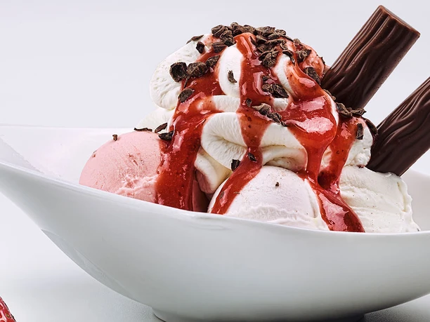 LaLuna Eine Eisschale mit Erdbeeren, Sahne, und Schokostäbchen, dekoriert mit roter Soße und Schokostreuseln.