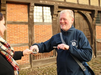 So schmeckt BZ.jpg Ein lächelnder Mann reicht einer Frau einen Ammerländer Löffeltrunk vor einem rustikalen Gebäude.