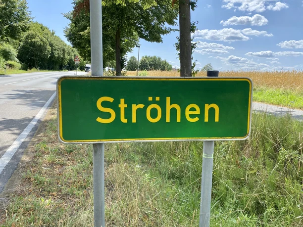 Ortseingangsschild von Ströhen, umgeben von Bäumen und Feldern unter blauem Himmel mit Wolken.