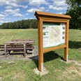 Holztafel mit Wanderkarte und Sitzbank auf einer Wiese unter blauem Himmel mit Wolken im Hintergrund.