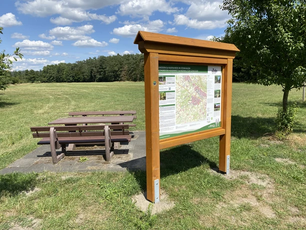 Holztafel mit Wanderkarte und Sitzbank auf einer Wiese unter blauem Himmel mit Wolken im Hintergrund.