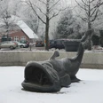 zwischenahner-wels-winter.jpg Bronzeskulptur des Zwischenahner Welses im Schnee, umgeben von winterlichen Bäumen und Häusern.