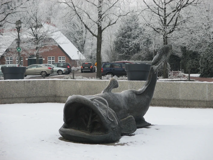 zwischenahner-wels-winter.jpg Bronzeskulptur des Zwischenahner Welses im Schnee, umgeben von winterlichen Bäumen und Häusern.