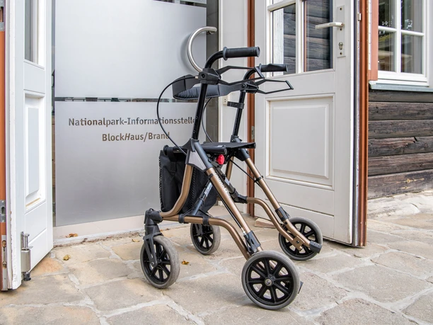 Barrièrevrije toegang tot het informatiecentrum Een bronskleurige rollator staat voor een open deur met het opschrift "National Park Information Point BlockHaus/Brandenberg".