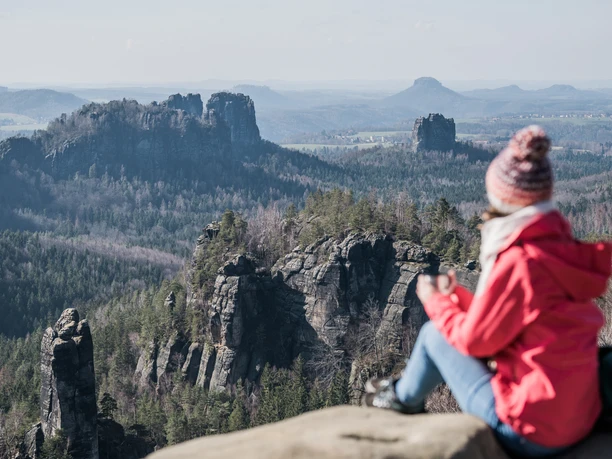 Winterliche Aussicht vom Carolafelsen Eine Person in roter Jacke und Mütze sitzt auf einem Felsen und blickt auf eine bewaldete Berglandschaft mit markanten Felsformationen.